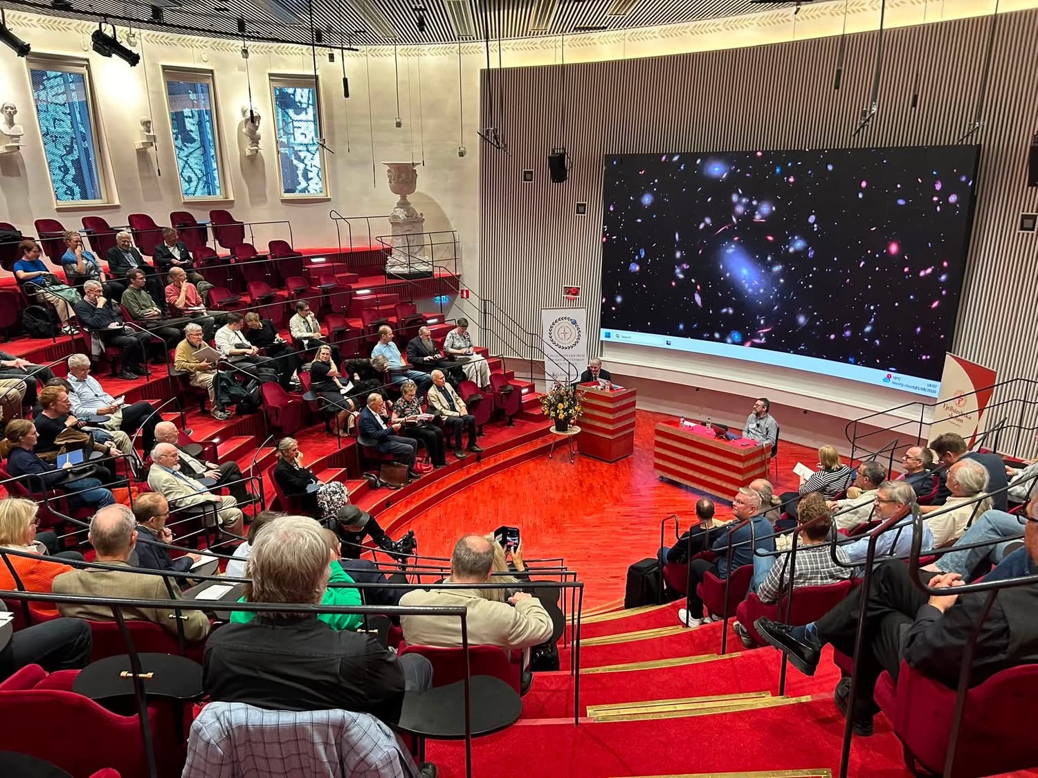 En publik som sitter i ett auditorium med en föreläsare längst ner. På skärmen bakom en flera galaxer.