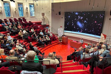 IMG_1359.jpeg En publik som sitter i ett auditorium med en föreläsare längst ner. På skärmen bakom en flera galaxer.