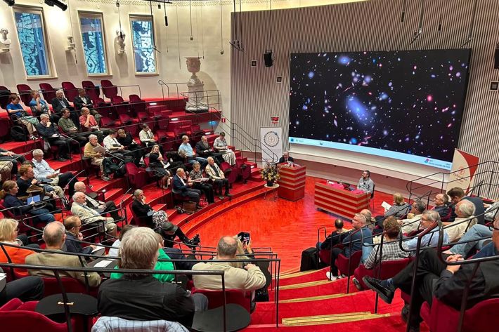 IMG_1359.jpeg En publik som sitter i ett auditorium med en föreläsare längst ner. På skärmen bakom en flera galaxer.