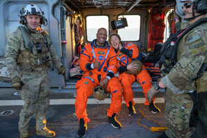 Victor Glover och Christina Koch.png Två glada astronauter i orangea rymddräkter sitter och tittar glada in i kameran. Bredvid dem står två militärer och ser lika glada ut.
