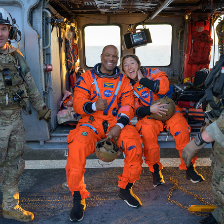 Victor Glover och Christina Koch.png Två glada astronauter i orangea rymddräkter sitter och tittar glada in i kameran. Bredvid dem står två militärer och ser lika glada ut.