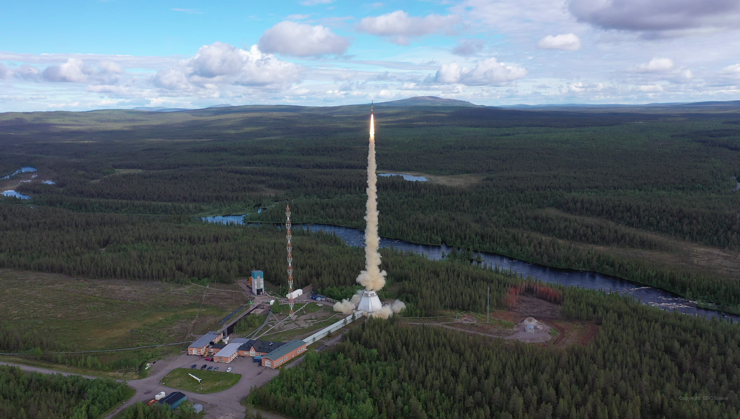 MASER14_vlcsnap-2019-07-04-09h33m24s373.png En raket lyfter från en rymdbas omgiven av granskog och älvar.