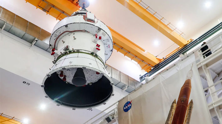 En bild tagen underifrån när rymdfarkosten Orion förflyttas till en vacuumkammare för testning med hjälp av en industriell lyftkran monterad i en stor industrilokal.