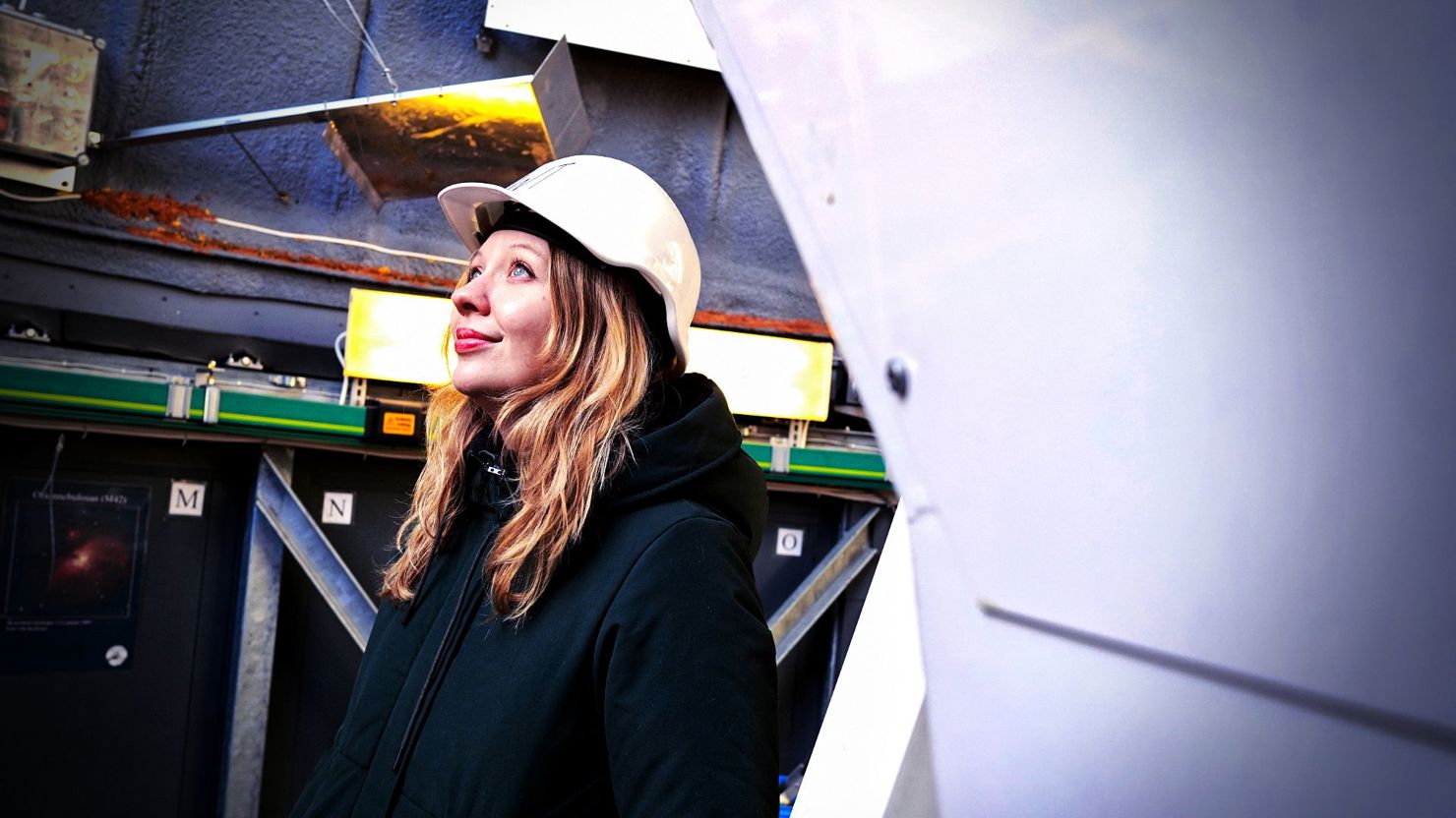 En kvinna med långt blont hår och läppstift (Linn Boldt-Christmas) bär skyddshjälm och mörk jacka. Hon står inomhus i ett teleskop och tittar uppåt med ett lugnt, förväntansfullt uttryck. Bakom henne syns teknisk utrustning, kablar och metallstrukturer.