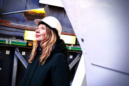 Linn Boldt-Christmas En kvinna med långt blont hår och läppstift (Linn Boldt-Christmas) bär skyddshjälm och mörk jacka. Hon står inomhus i ett teleskop och tittar uppåt med ett lugnt, förväntansfullt uttryck. Bakom henne syns teknisk utrustning, kablar och metallstrukturer.