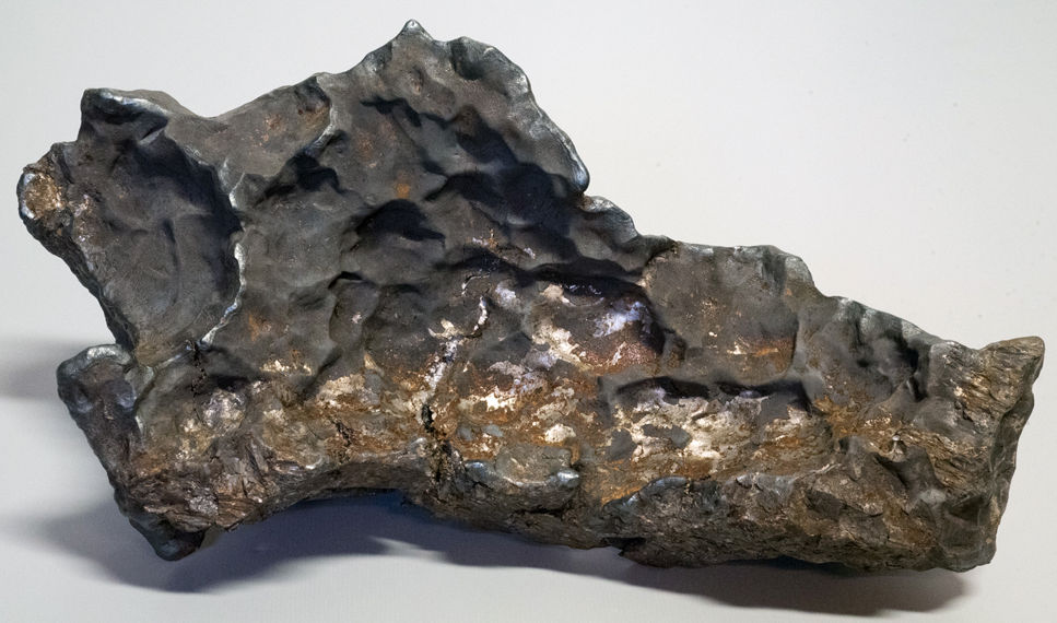 Den ojämna, mörkgråa meteoriten med metalliskt skimmer och gropar i ytan, liggande på ett ljust underlag.