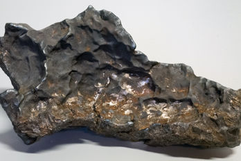 Meteoriten i Enköping Den ojämna, mörkgråa meteoriten med metalliskt skimmer och gropar i ytan, liggande på ett ljust underlag.