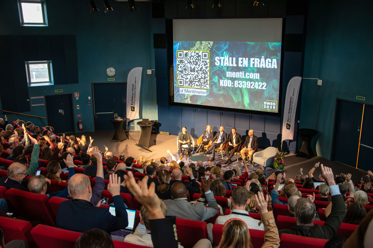 En publik räcker upp händerna till en intresserad panel.