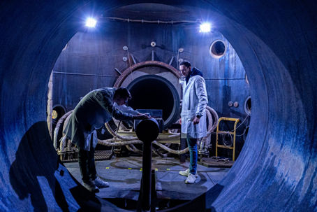 Team_preparing_the_plasma_wind_tunnel.jpg Två personer som står i en halvt upplyst tunnel och förbereder för ett sorts test.